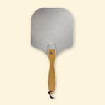 Pelle à pizza en aluminium vue de face, avec large plateau rectangulaire arrondi et manche en bois équipé d’une lanière de suspension.