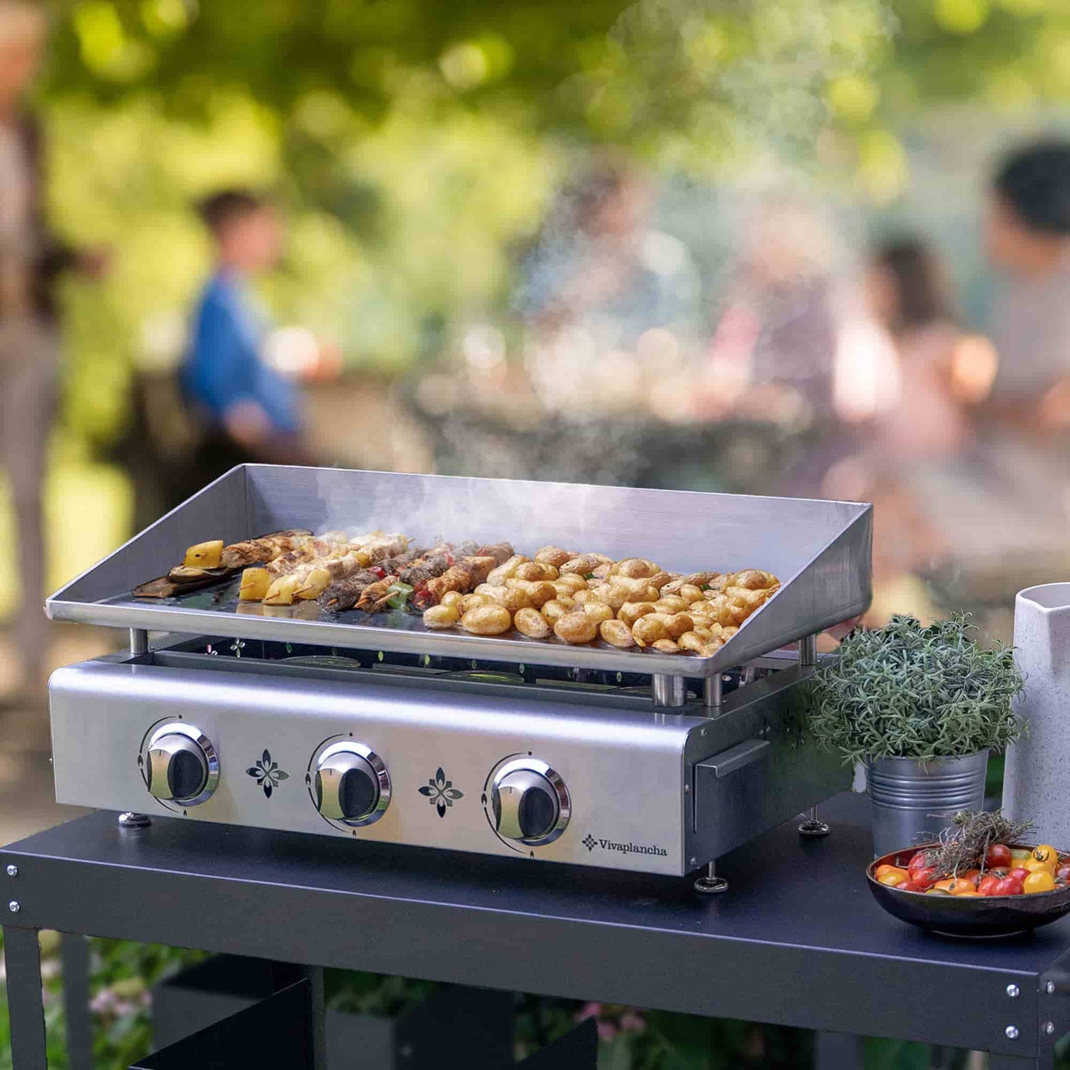 Plancha Vivaplancha en inox en cours de cuisson avec brochettes et pommes de terre, sur un chariot extérieur, avec de la fumée qui s’élève et un groupe de personnes floues en arrière-plan dans un jardin.