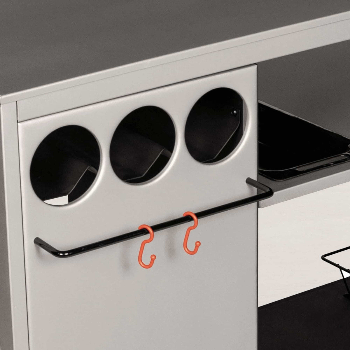 Module de cuisine extérieure avec trois rangements cylindriques intégrés et une barre porte-ustensiles noire équipée de crochets orange, présenté en gros plan.