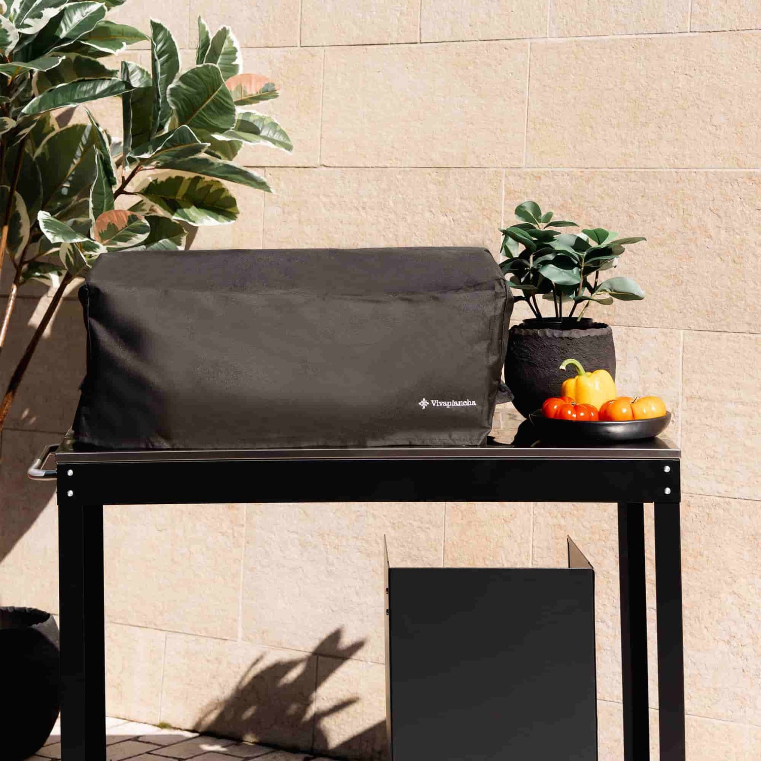 Housse noire de protection pour plancha deux brûleurs Vivaplancha, posée sur une desserte en extérieur, entourée de plantes et de légumes, présentée en situation réelle.