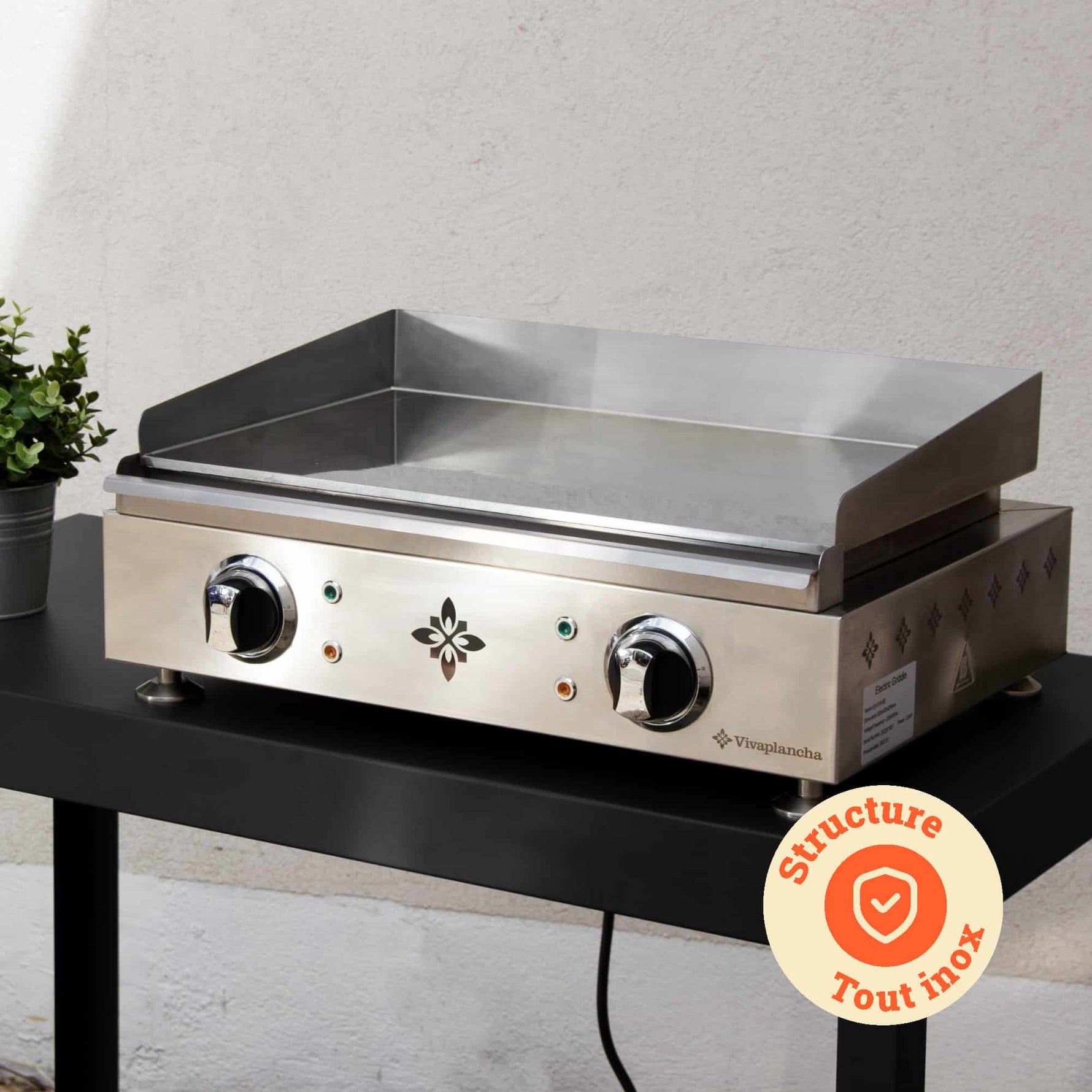 Vivaplancha-Plancha auf einem Außentisch platziert, mit ihrer vollständig aus Edelstahl gefertigten Struktur im Vordergrund und einem „Struktur komplett aus Edelstahl“-Abzeichen, das in einem orangefarbenen Kreis angezeigt wird.