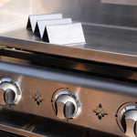 Supports de maintien au chaud en inox posés sur la plaque d’une plancha inox à trois brûleurs, présentés en gros plan en extérieur.